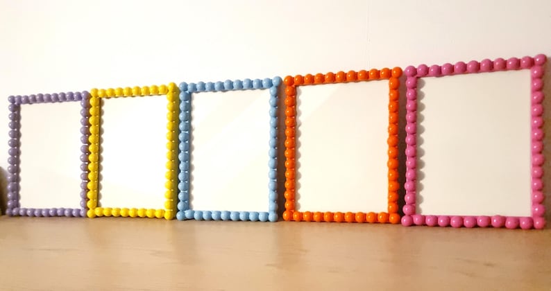 May include: Five colorful picture frames with white interiors. The frames are arranged in a row and feature a beaded border in purple, yellow, light blue, orange, and pink. The frames are on a wooden surface.