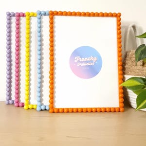 May include: Four colorful picture frames with beaded edges. The frames are in shades of purple, pink, yellow, blue, and orange. Each frame has a white interior. The orange frame has a sticker that says "Francky Paillettes".
