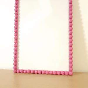 May include: A rectangular pink frame with a beaded border. The frame is empty, with a white interior. The frame is sitting on a light brown surface, against a white wall.
