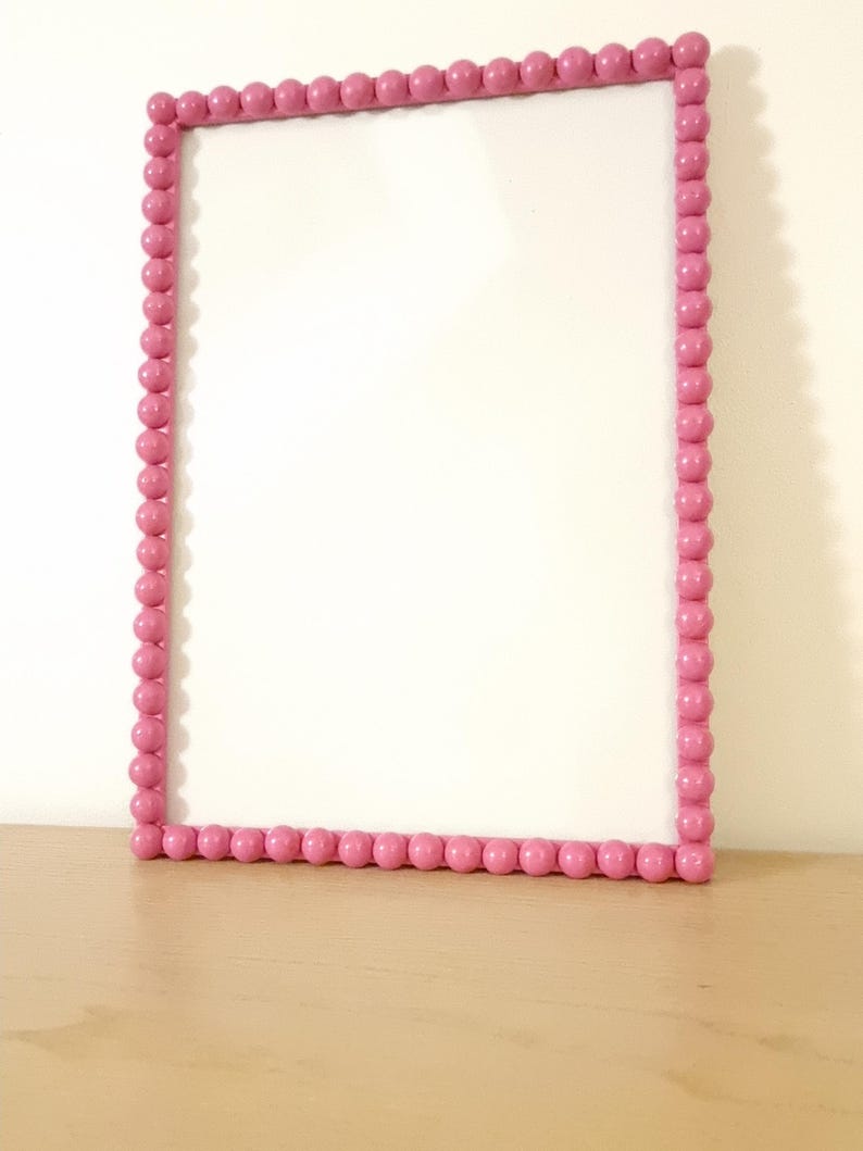 May include: A rectangular pink frame with a beaded border. The frame is empty, with a white interior. The frame is sitting on a light brown surface, against a white wall.