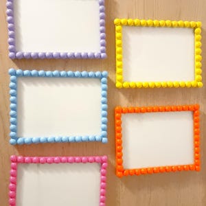 May include: Five rectangular picture frames with white centers and colorful beaded borders. The frames are in shades of purple, yellow, blue, orange, and pink. The frames are mounted on a light brown wooden surface.