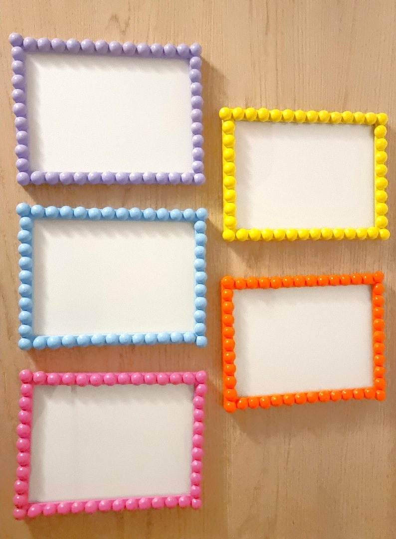 May include: Five rectangular picture frames with white centers and colorful beaded borders. The frames are in shades of purple, yellow, blue, orange, and pink. The frames are mounted on a light brown wooden surface.