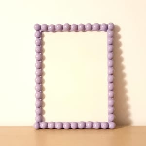 May include: A rectangular picture frame with a light purple beaded border. The frame is empty, with a white interior. The frame is sitting on a light brown surface, with a white wall in the background.