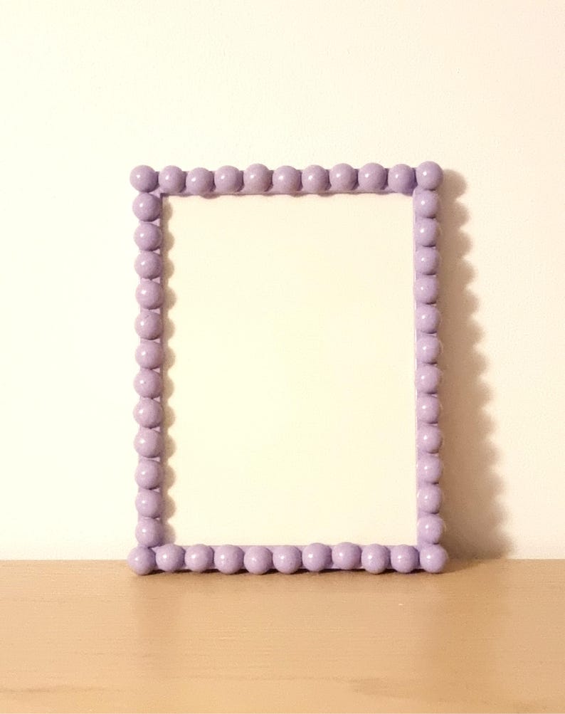 May include: A rectangular picture frame with a light purple beaded border. The frame is empty, with a white interior. The frame is sitting on a light brown surface, with a white wall in the background.