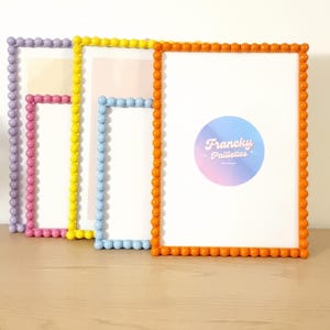 May include: Four colorful picture frames with beaded edges. The frames are in shades of purple, pink, yellow, light blue, and orange. The frames are arranged on a light brown wooden surface. The rightmost frame has a circular logo with the text "Francky Paillettes".