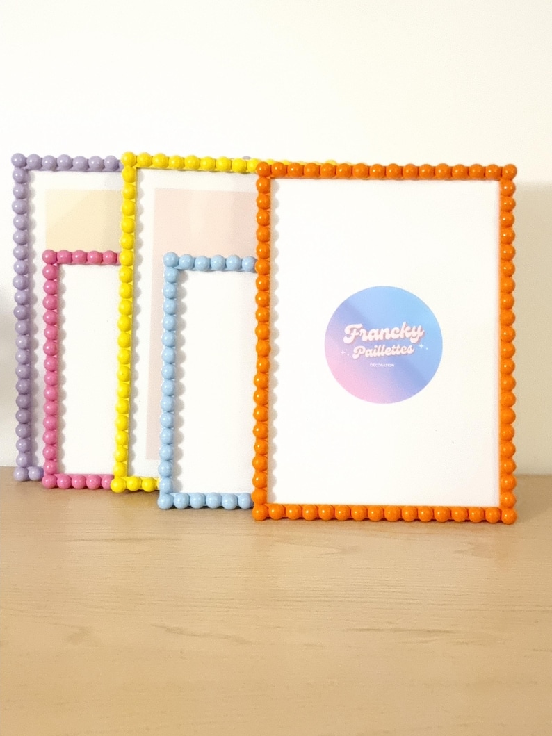 May include: Four colorful picture frames with beaded edges. The frames are in shades of purple, pink, yellow, light blue, and orange. The frames are arranged on a light brown wooden surface. The rightmost frame has a circular logo with the text "Francky Paillettes".