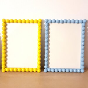 May include: Two rectangular picture frames. One frame is yellow, the other is light blue. Both frames have a white interior and are decorated with a border of small, round, raised beads. The frames are displayed on a wooden surface.