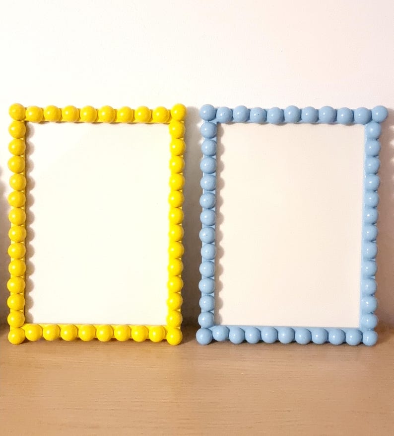 May include: Two rectangular picture frames. One frame is yellow, the other is light blue. Both frames have a white interior and are decorated with a border of small, round, raised beads. The frames are displayed on a wooden surface.