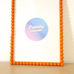 May include: An orange beaded picture frame with a white interior. The frame has a rectangular shape and is decorated with small, round beads. A circular logo with the text "Francky Paillettes" is visible inside the frame.