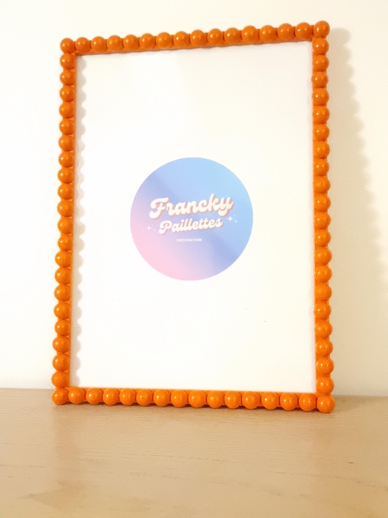May include: An orange beaded picture frame with a white interior. The frame has a rectangular shape and is decorated with small, round beads. A circular logo with the text "Francky Paillettes" is visible inside the frame.