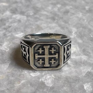 Puede incluir: Un anillo de plata con una cara cuadrada que presenta un diseño de cruz de Jerusalén. El anillo tiene un acabado pulido y un patrón detallado en los lados. El anillo está sobre una superficie gris texturizada.