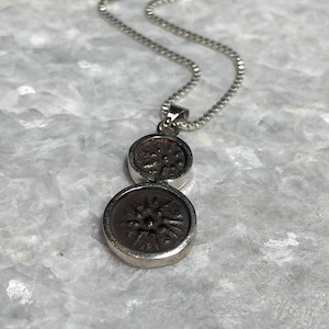 Op de afbeelding: Zilverkleurige ketting met een hanger met twee cirkelvormige, donkerbruine medaillons. Elk medaillon heeft een verhoogd bloemmotief. De ketting is een delicate, vierkante schakelketting, tegen een lichte, gemarmerde achtergrond.