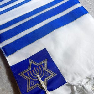 Peut inclure: Châle de prière à rayures blanches et bleues avec un carré bleu orné d'une étoile de David et d'un design de menorah dorés. Le châle a des glands blancs et est fait d'un tissu doux et fluide.