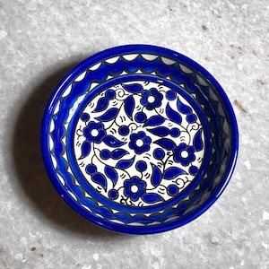 Puede incluir: Un cuenco redondo de cerámica pintado a mano con un vibrante diseño floral azul y blanco. El cuenco presenta un borde azul oscuro y un patrón detallado de hojas y flores azules sobre un fondo blanco. El cuenco tiene unos 15 cm de diámetro.