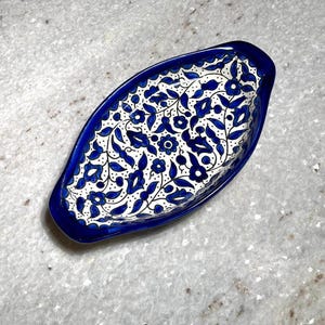 May include: Oval-shaped ceramic dish with a vibrant blue floral pattern on a white background. The dish has a deep blue rim and is likely used for serving or display. The design features stylized flowers and leaves.