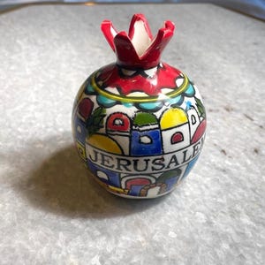 Puede incluir: Un objeto decorativo de cerámica con forma de granada. La parte superior es roja con un diseño en forma de corona. El cuerpo está pintado con un paisaje urbano colorido y la palabra "JERUSALEM" en azul. El artículo está sobre una superficie clara.