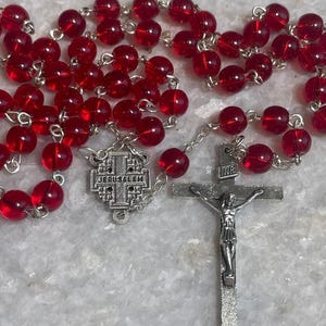 May include: A red rosary with round beads and silver-toned metal accents. The rosary features a crucifix with the inscription "INRI" and a Jerusalem emblem. The beads are a deep, translucent red.
