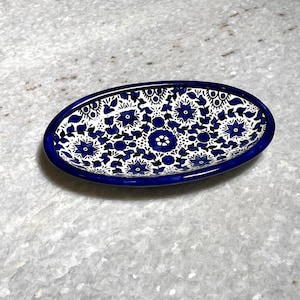 Puede incluir: Plato ovalado de cerámica con un vibrante estampado floral azul y blanco. El plato tiene un borde azul oscuro y un acabado brillante. El diseño floral presenta flores y hojas estilizadas, creando una pieza decorativa y artística.