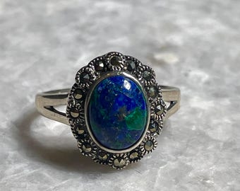 May include: A silver ring featuring an oval-shaped, blue and green gemstone. The gemstone is surrounded by a decorative border of smaller, dark stones. The ring's band is a simple, split design.
