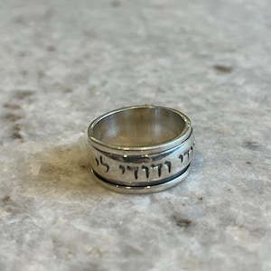 Puede incluir: Un anillo de plata con texto hebreo grabado alrededor de la banda. El anillo tiene una banda ancha y un acabado pulido. El anillo está sobre un fondo neutro.