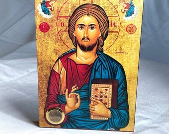May include: A religious icon featuring a depiction of Jesus Christ. The image shows Jesus with a beard, holding a book, and wearing a blue robe and red tunic. The background is gold with red accents and two angels.
