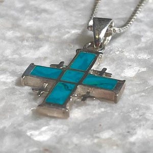Może przedstawiać: Srebrny wisiorek w kształcie krzyża z inkrustacją z turkusu, zawieszony na delikatnym srebrnym łańcuszku. Krzyż ma klasyczny design z prostokątnymi kamieniami z turkusu osadzonymi w srebrnej ramie. Wisiorek jest prezentowany na teksturowanej, jasnej powierzchni.