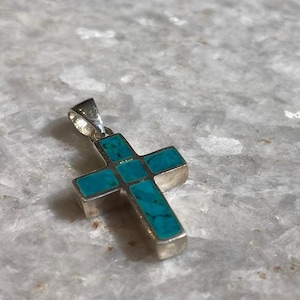 Puede incluir: Colgante de cruz de plata con incrustaciones de turquesa. La cruz presenta un diseño clásico con una piedra de turquesa rectangular en cada sección. El colgante tiene un pequeño lazo en la parte superior para una cadena.