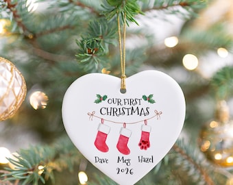 May include: A white heart-shaped ceramic ornament hanging by a gold string. It features the text "OUR FIRST CHRISTMAS" above an illustration of three red stockings, with the names "Dave", "Meg", and "Hazel" and the year "2026" printed below.