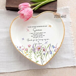 May include: A white heart-shaped ceramic trinket dish with a gold-toned rim, featuring a floral print at the bottom and text that reads: TO MY FUTURE DAUGHTER-IN-LAW Serena I may not have given you the gift of life. But life has given me the gift of you. Thank you for loving my son. Love, Gina.