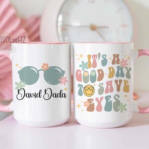 May include: Two white ceramic mugs with pink handles and rims. One mug features sunglasses and the name "David Dada." The other mug has the text "It's a good day to say yes" with a smiley face.