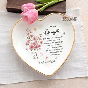 May include: A white heart-shaped ceramic trinket dish with a gold-painted rim. The dish features a pink floral illustration, small butterflies, and the text: To our Daughter, Even when we're not close by, we want you to know we love you for the little girl that you once were, for the amazing woman you are today, and for the precious daughter you will always be. Love, Mom & Dad. The name Nicole is printed vertically along the floral stem.