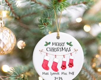 May include: A white ceramic Christmas ornament with a gold string, featuring the text "MERRY CHRISTMAS" and three red stockings hanging from a string. The names "Matt Lynnett Max Puff" and the year "2025" are also printed on the ornament.