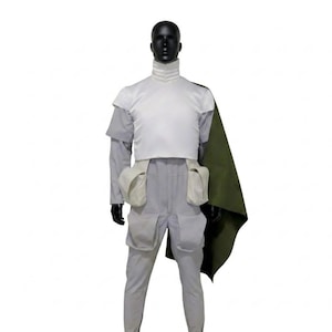 May include: A costume displayed on a black mannequin. The outfit comprises a white top, grey trousers, and a green cape. The top features a high collar and a white vest. The trousers have two large thigh pockets. The cape is draped over the shoulder.