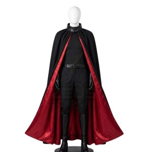 Puede incluir: Una capa negra de cuerpo entero con forro rojo vibrante, sobre una camisa negra de manga larga, pantalones negros con cinturón negro y botas negras. El conjunto se exhibe en un maniquí blanco, con cuello alto y guantes negros.