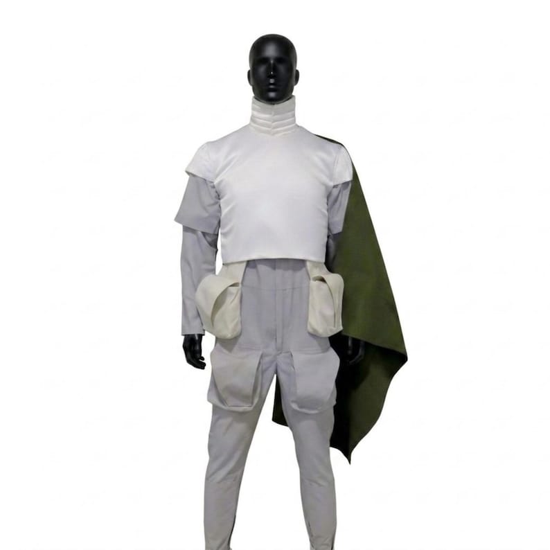 May include: A costume displayed on a black mannequin. The outfit comprises a white top, grey trousers, and a green cape. The top features a high collar and a white vest. The trousers have two large thigh pockets. The cape is draped over the shoulder.