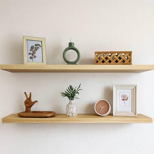 May include: Two light wood floating shelves against a white wall. The top shelf holds a framed picture, a green candle holder, and a wooden box. The bottom shelf has a wooden hand sculpture, a vase, a clock, and a framed picture.