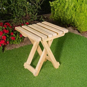 Könnte beinhalten: Kleiner, hellfarbiger, klappbarer Beistelltisch aus Holz mit Lattenrost. Der Tisch steht auf einem grünen Rasen, im Hintergrund rote Blumen und grüne Büsche. Der Tisch ist etwa 46 cm lang.