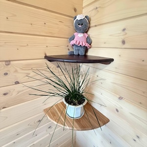 Può includere: Una bambola di gatto di peluche grigia con un vestito rosa e una fascia bianca è seduta su una mensola angolare marrone scuro. Sotto, una pianta in vaso con lunghe foglie verdi poggia su una mensola angolare in legno chiaro. Le mensole sono montate su una parete a pannelli di legno chiaro.