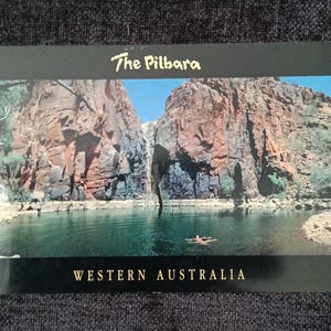 Op de afbeelding: Een ansichtkaart met een schilderachtig uitzicht op The Pilbara in West-Australië. De afbeelding toont een waterpartij omgeven door grote, roodbruine rotsformaties onder een heldere blauwe lucht. De tekst "The Pilbara" en "WESTERN AUSTRALIA" staan op de kaart.