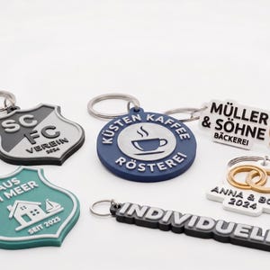 May include: A collection of five custom rubber keychains featuring various designs: a shield-shaped SC FC VEREIN 2024, a round KÜSTEN KAFFEE RÖSTEREI, a MÜLLER & SÖHNE BÄCKEREI with a pretzel, a shield-shaped HAUS AM MEER SEIT 2023, and a rectangular INDIVIDUELL.