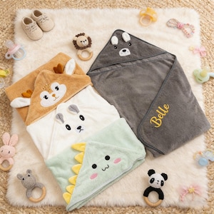 May include: A collection of baby hooded towels in various colors, including brown, white, and green, each with an animal face design. The towels are surrounded by baby accessories like pacifiers, shoes, and teethers.