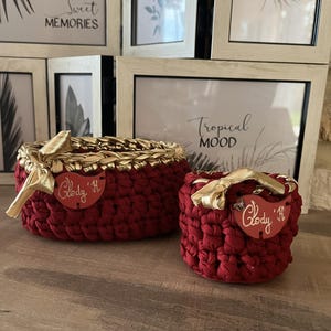 Peut inclure: Deux paniers en crochet rouges faits à la main avec des bordures et des accents dorés. Le plus grand panier a une forme ovale, tandis que le plus petit est rond. Les deux ont un nœud doré et une étiquette en forme de cœur avec le nom "Glody 'H".