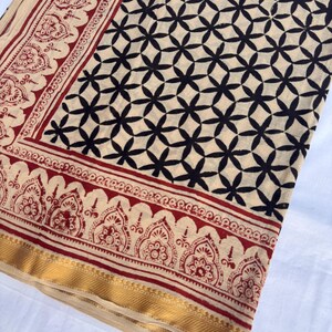 May include: A textile fabric featuring a cream-colored base with a repeating black geometric floral pattern. The border displays a red and cream intricate paisley-style motif, finished with a solid gold-toned band along the bottom edge.