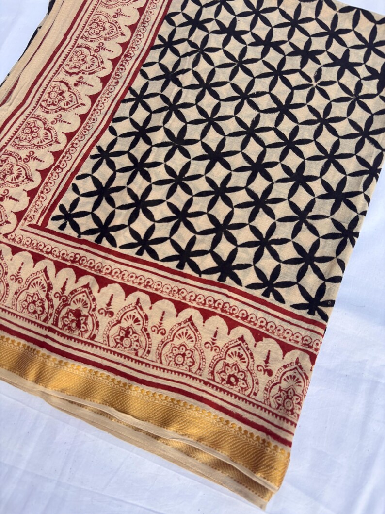 May include: A textile fabric featuring a cream-colored base with a repeating black geometric floral pattern. The border displays a red and cream intricate paisley-style motif, finished with a solid gold-toned band along the bottom edge.