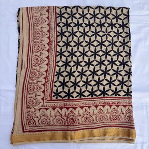 May include: A rectangular textile featuring a beige base with a black geometric floral pattern across the main body. The border displays a dark red intricate paisley-style print, finished with a solid gold-toned band along the bottom edge.