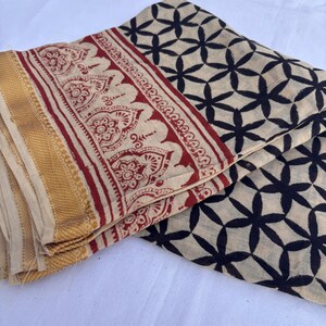 May include: A folded textile featuring a cream-colored base with a black repeating floral pattern. The fabric includes a wide border with a red intricate botanical print and a solid gold-toned textured edge along the side of the material.