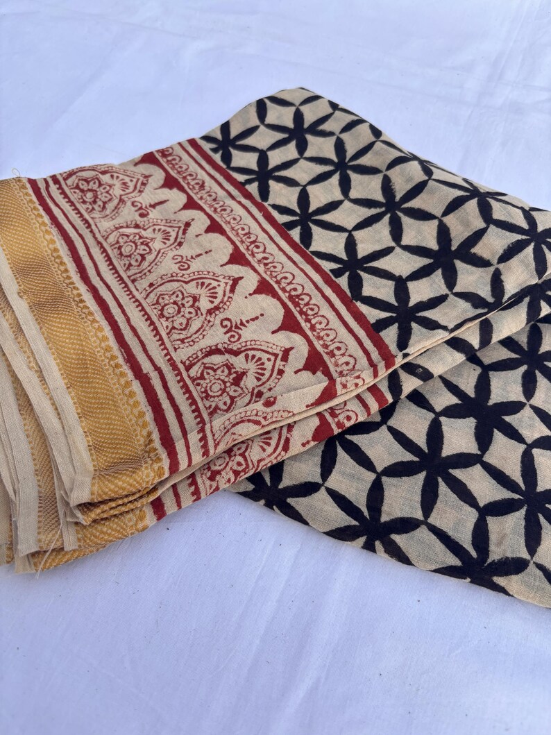 May include: A folded textile featuring a cream-colored base with a black repeating floral pattern. The fabric includes a wide border with a red intricate botanical print and a solid gold-toned textured edge along the side of the material.