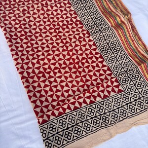 May include: A textile fabric featuring a repeating geometric pattern of red triangles on a cream background. The border displays a black floral motif followed by thin stripes in yellow, green, and red, finished with a plain cream-colored edge.