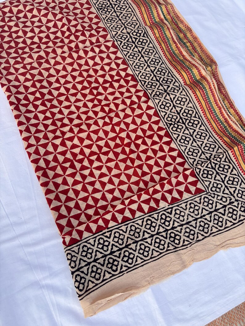 May include: A textile fabric featuring a repeating geometric pattern of red triangles on a cream background. The border displays a black floral motif followed by thin stripes in yellow, green, and red, finished with a plain cream-colored edge.