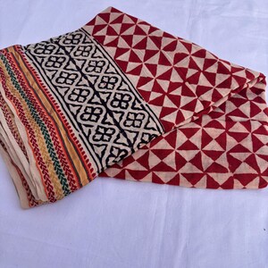 May include: A folded textile featuring a repeating red and cream triangular geometric pattern. The border displays a black floral motif on a cream background, finished with thin, parallel stripes in red, green, and yellow along the edge.